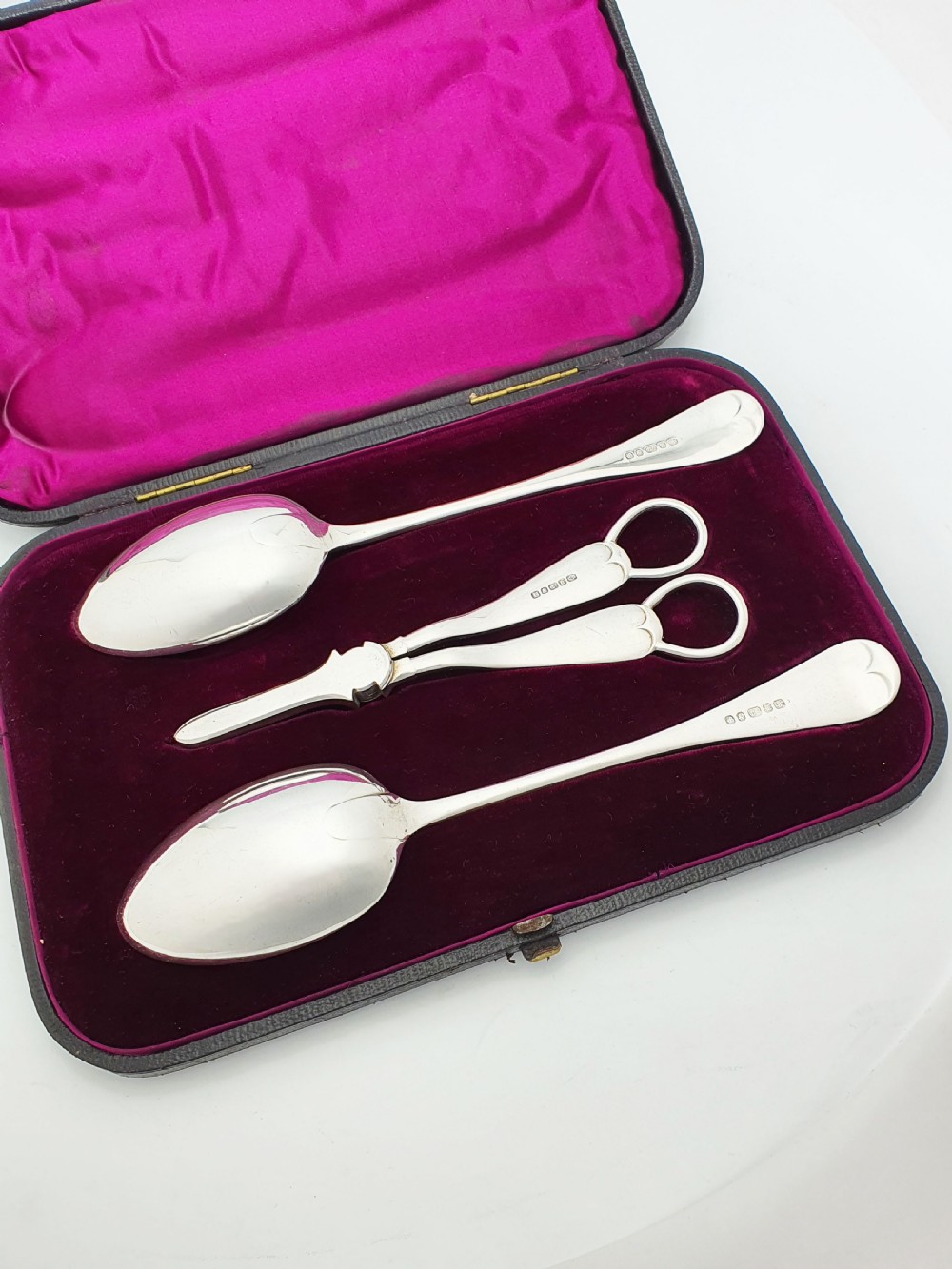 An open black presentation box lined with magenta velvet containing two engraved silver serving spoons and matching grape shears. View VI
