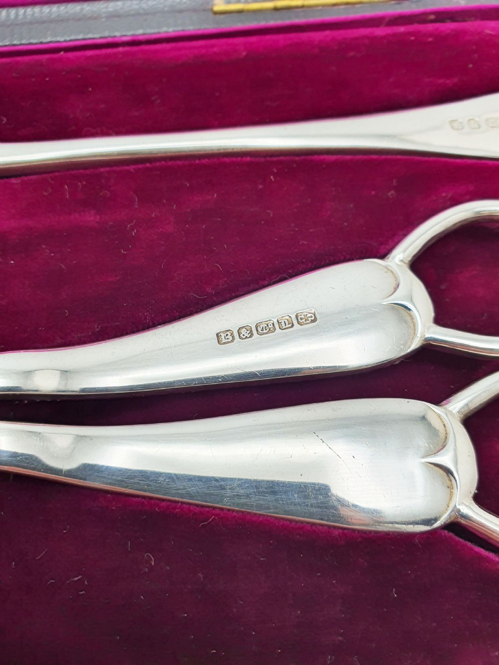 An open black presentation box lined with magenta velvet containing two engraved silver serving spoons and matching grape shears. View VII