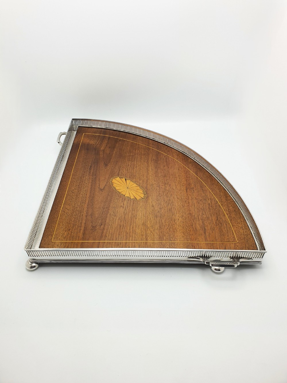 A fan-shaped Edwardian mahogany serving tray featuring an inlaid oval patera motif, fine line stringing, and a pierced silver-plated gallery rim with handles and ball feet.