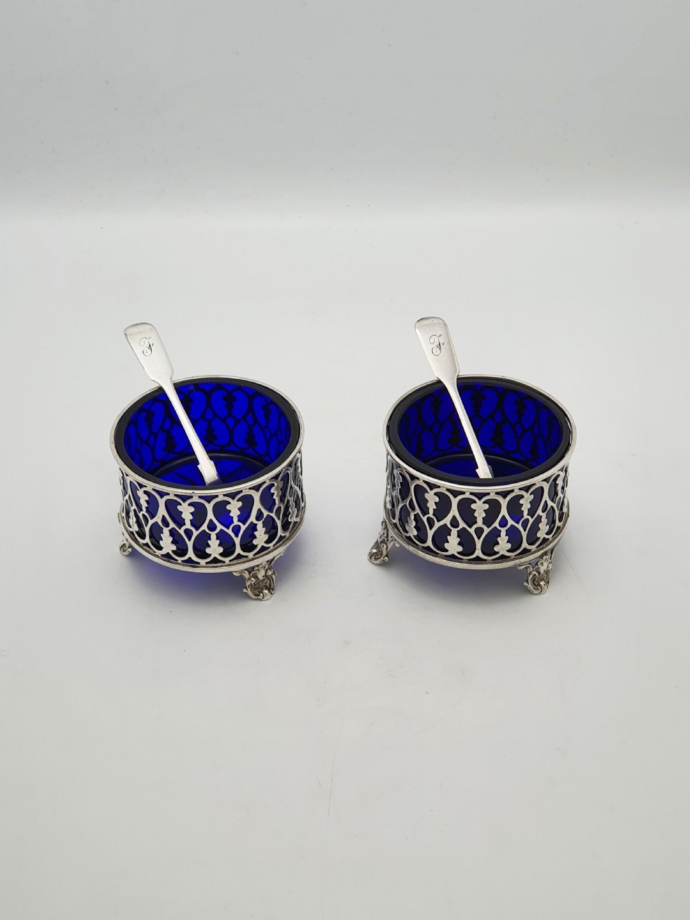 A pair of circular pierced sterling silver salt cellars containing dark cobalt blue glass liners with accompanying engraved silver spoons.