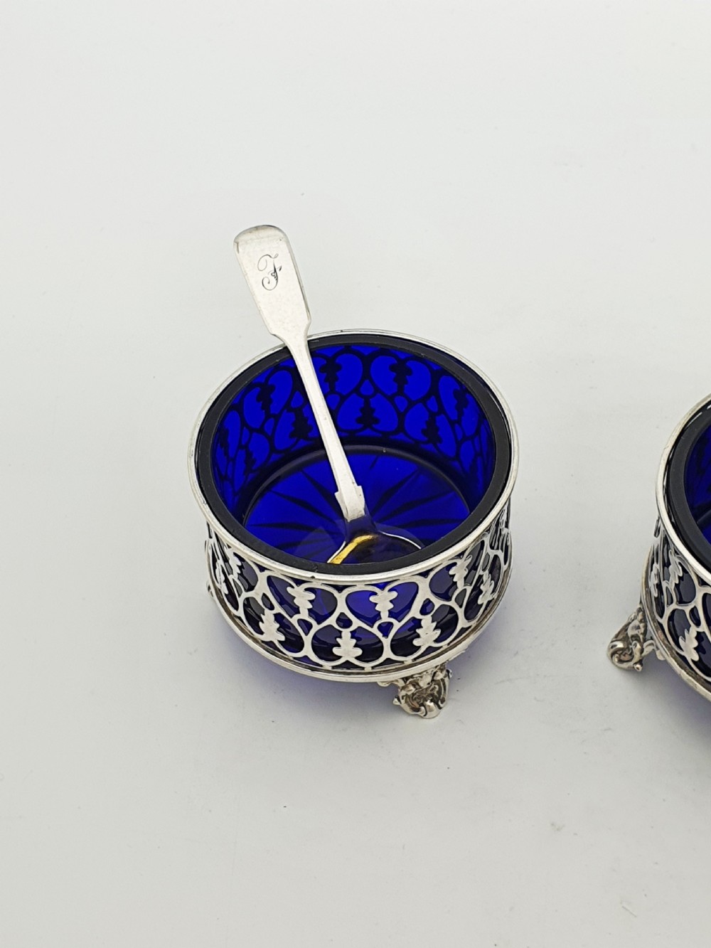 A pair of circular pierced sterling silver salt cellars containing dark cobalt blue glass liners with accompanying engraved silver spoons. View II