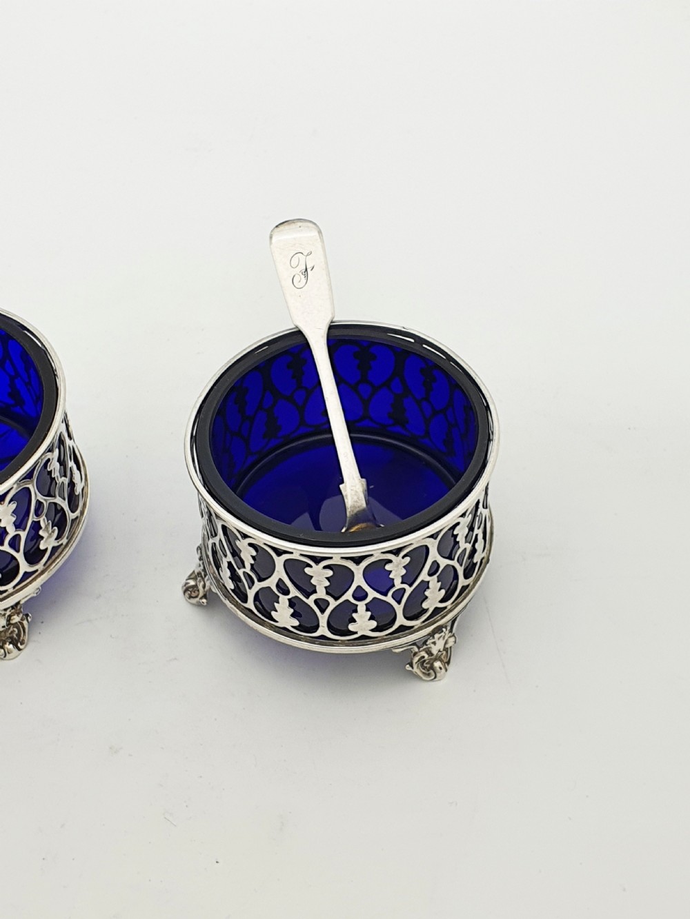 A pair of circular pierced sterling silver salt cellars containing dark cobalt blue glass liners with accompanying engraved silver spoons. View III