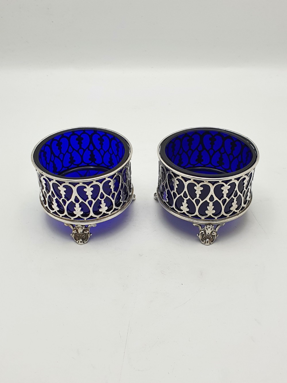 A pair of circular pierced sterling silver salt cellars containing dark cobalt blue glass liners with accompanying engraved silver spoons. View IV