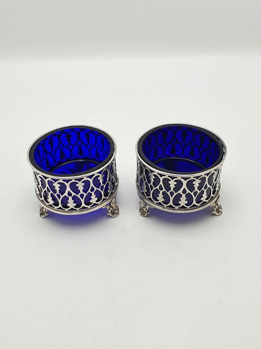 A pair of circular pierced sterling silver salt cellars containing dark cobalt blue glass liners with accompanying engraved silver spoons. View V