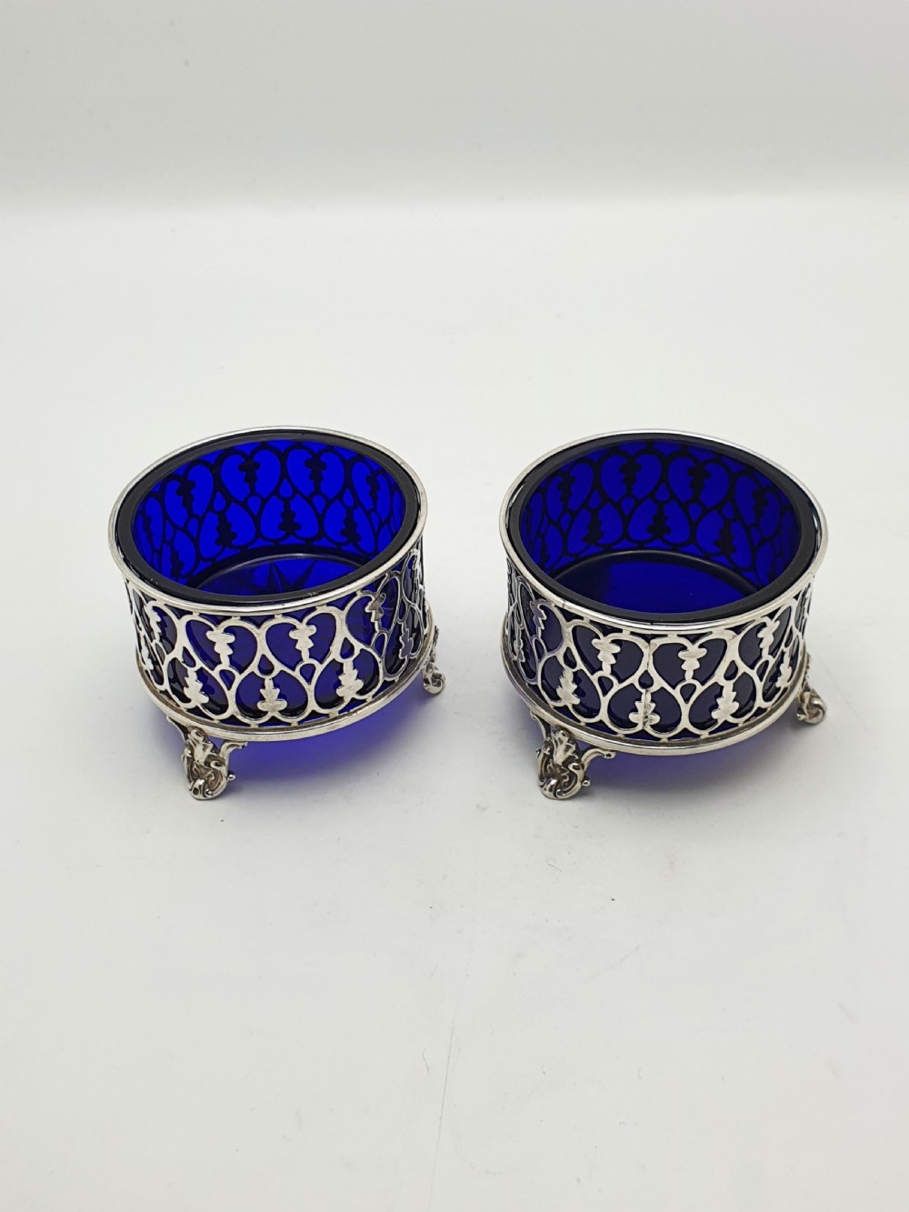 A pair of circular pierced sterling silver salt cellars containing dark cobalt blue glass liners with accompanying engraved silver spoons. View VI