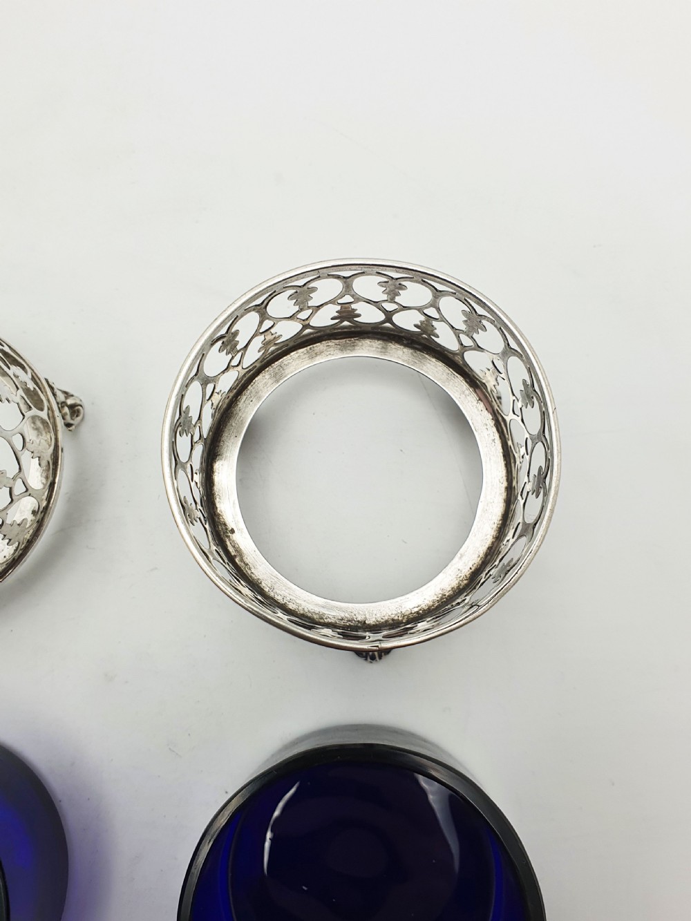 A pair of circular pierced sterling silver salt cellars containing dark cobalt blue glass liners with accompanying engraved silver spoons. View IX
