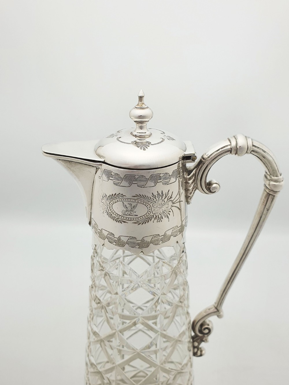 A tall Victorian cut glass claret jug featuring geometric diamond-cut motifs and an engraved silver-plated mount with a hinged lid and scrolled handle. View VII