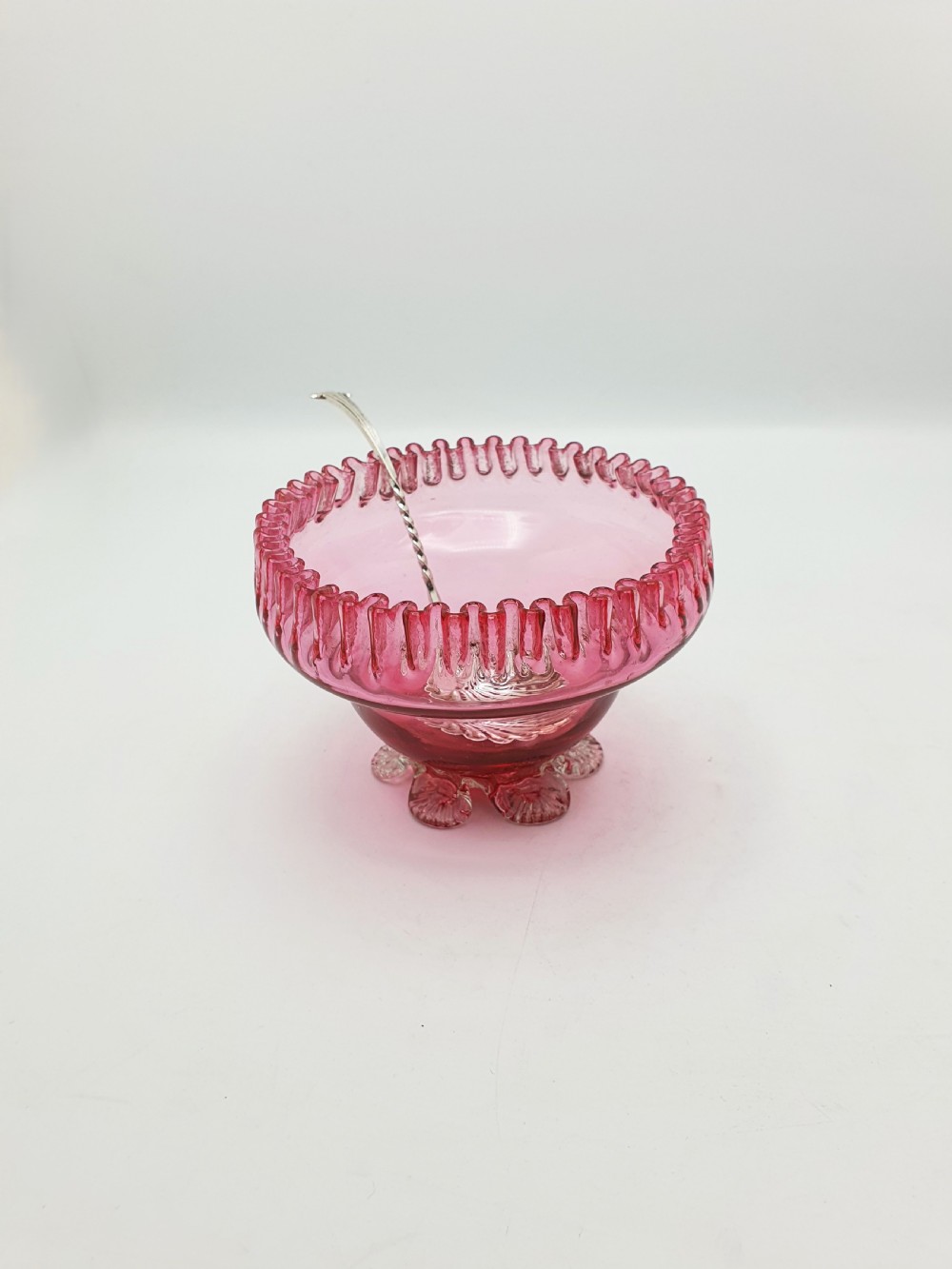 A Victorian cranberry glass bowl with a crimped rim and applied feet, containing a metal pierced spoon.