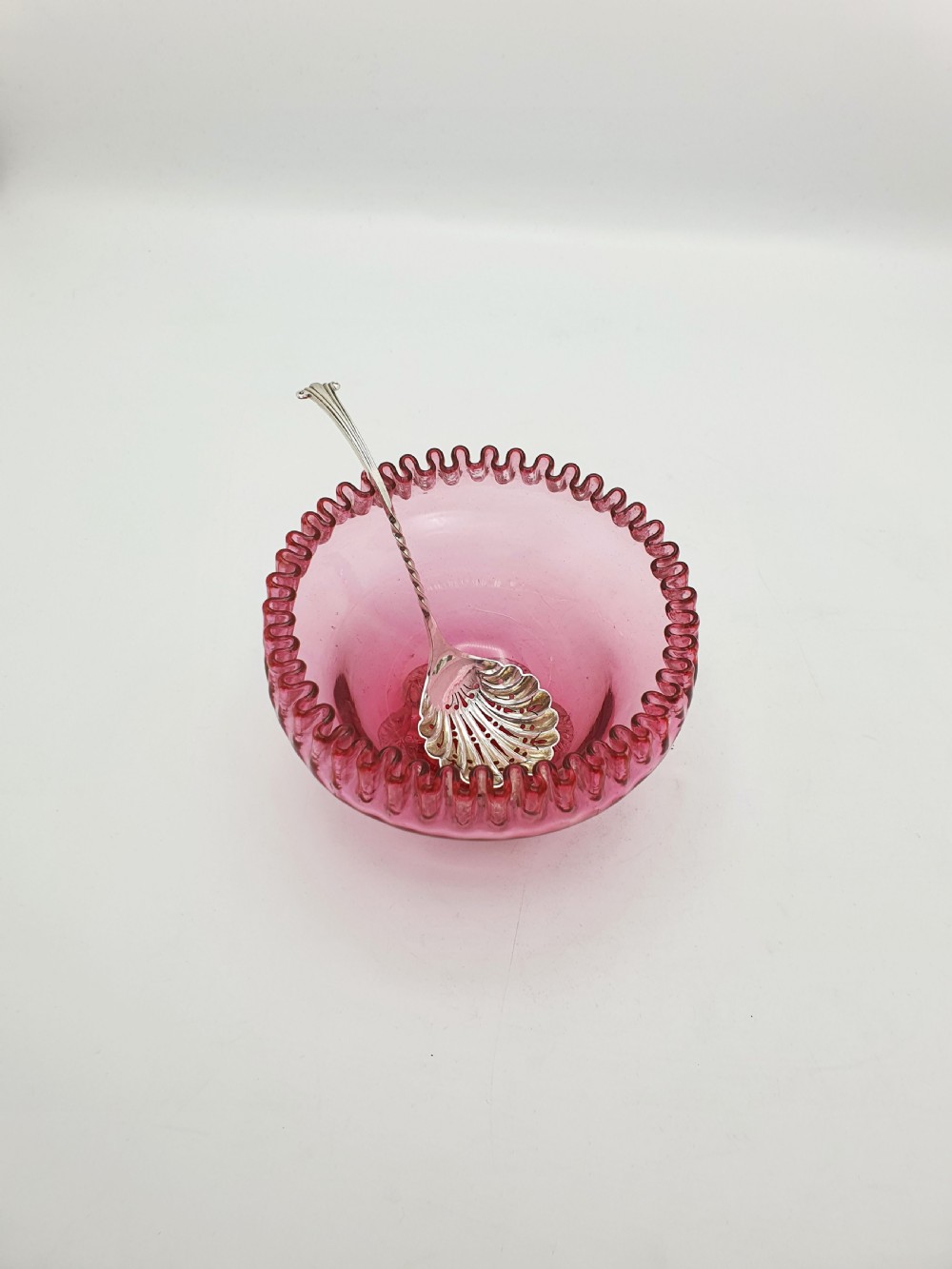 A Victorian cranberry glass bowl with a crimped rim and applied feet, containing a metal pierced spoon. View II