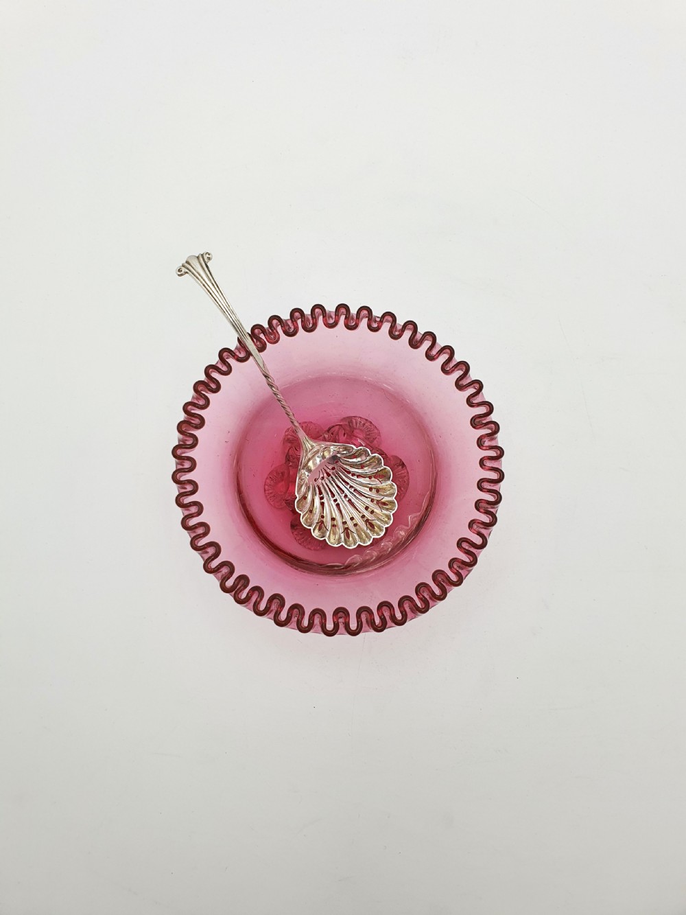 A Victorian cranberry glass bowl with a crimped rim and applied feet, containing a metal pierced spoon. View III