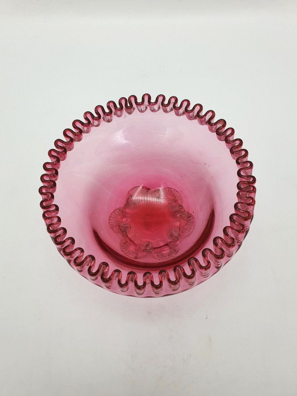 A Victorian cranberry glass bowl with a crimped rim and applied feet, containing a metal pierced spoon. View IV