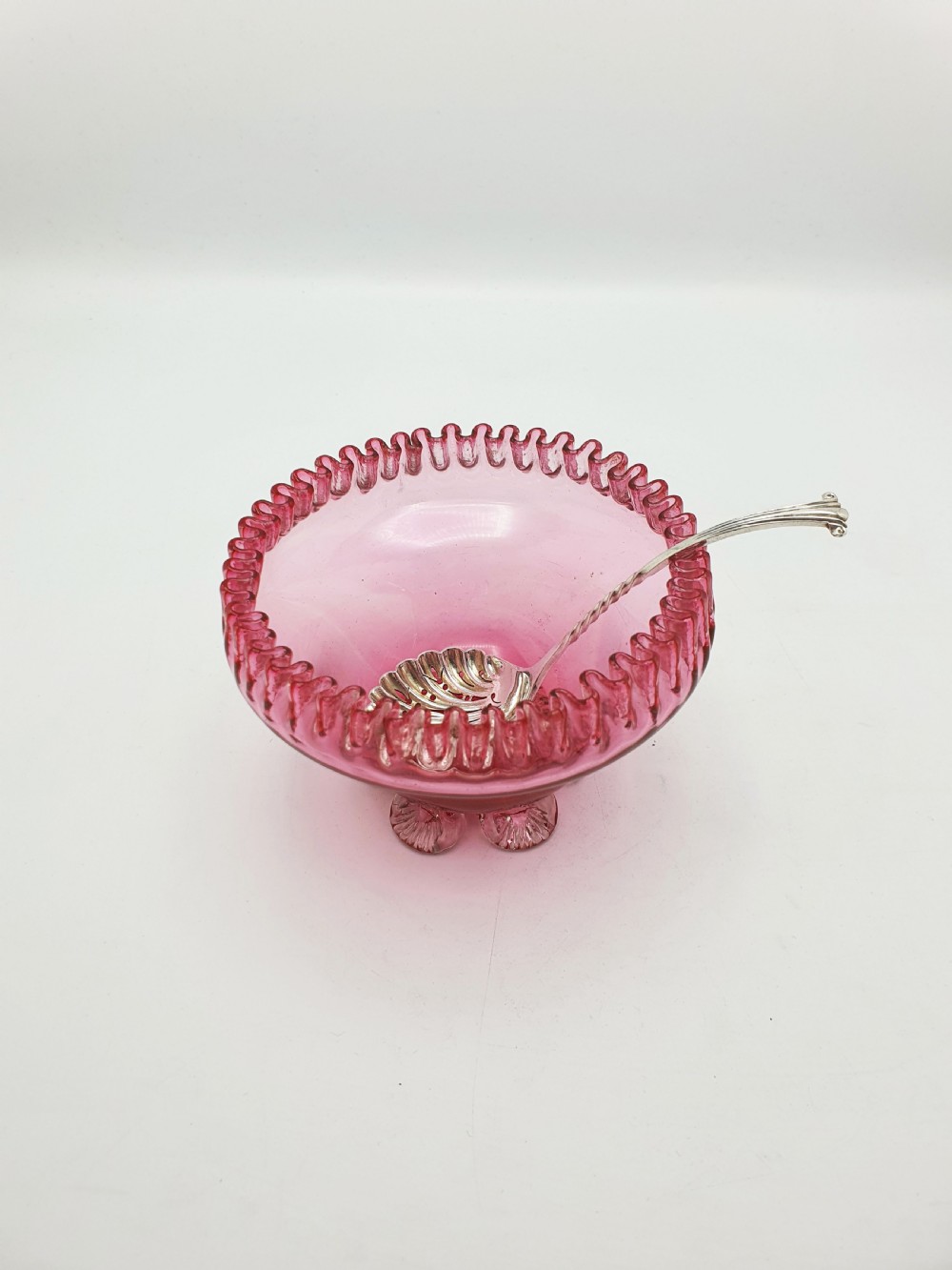 A Victorian cranberry glass bowl with a crimped rim and applied feet, containing a metal pierced spoon. View VII
