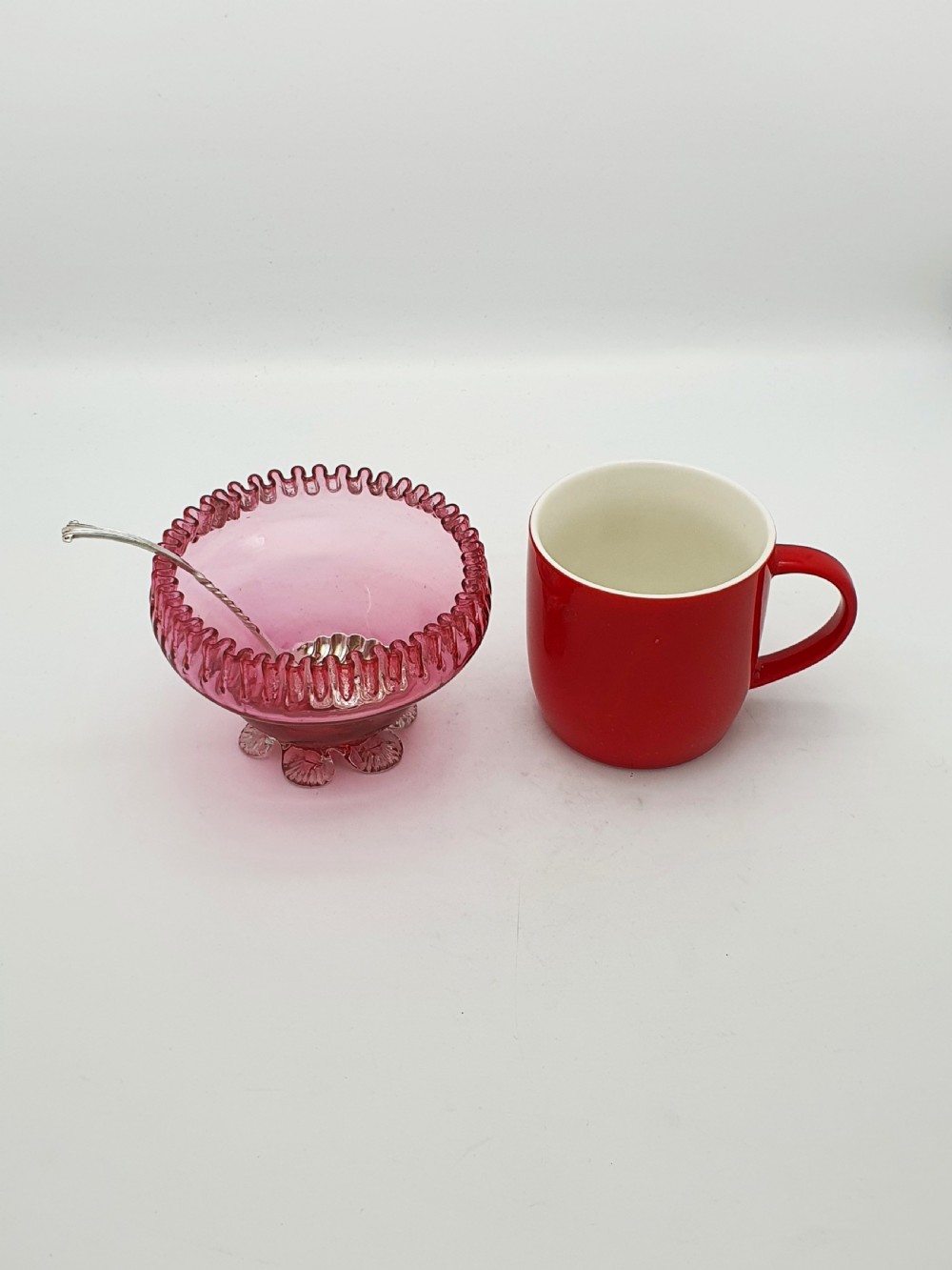 A Victorian cranberry glass bowl with a crimped rim and applied feet, containing a metal pierced spoon. View IX