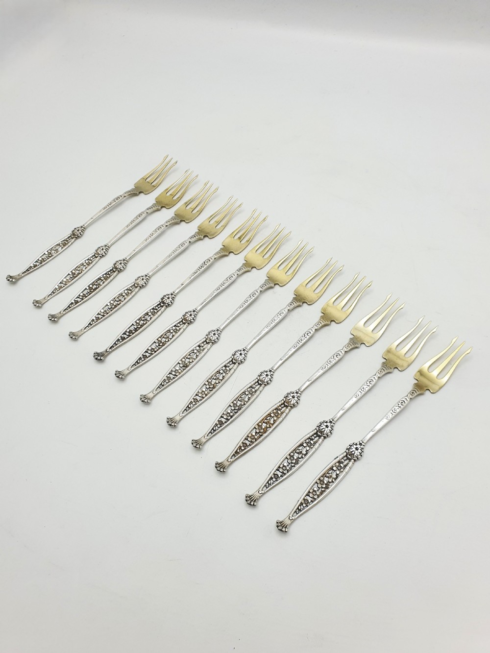 A set of twelve sterling silver seafood forks with ornate floral handles and gold gilt tines arranged in a diagonal line.