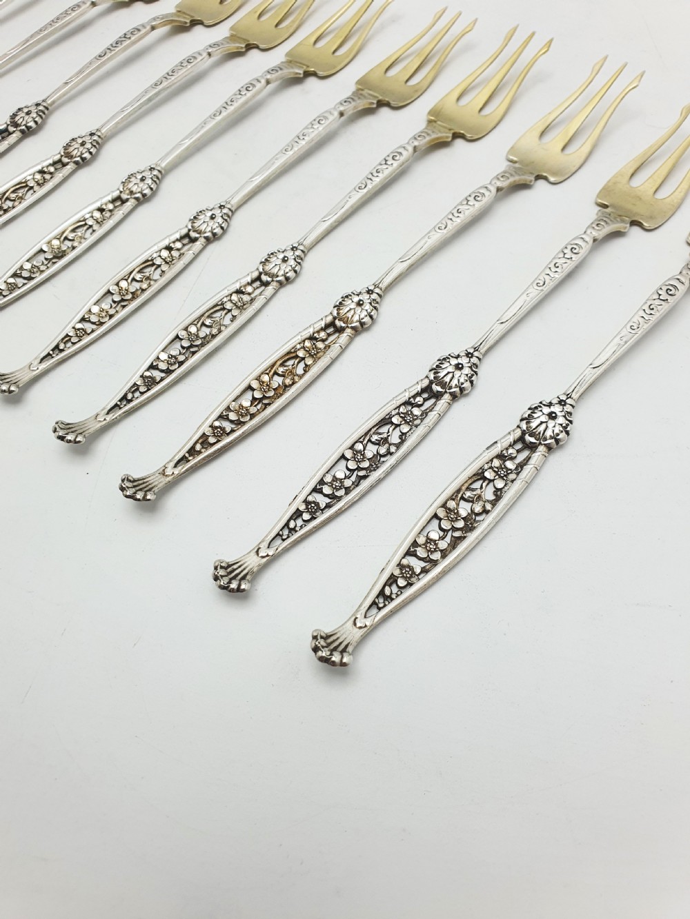 A set of twelve sterling silver seafood forks with ornate floral handles and gold gilt tines arranged in a diagonal line. View IV