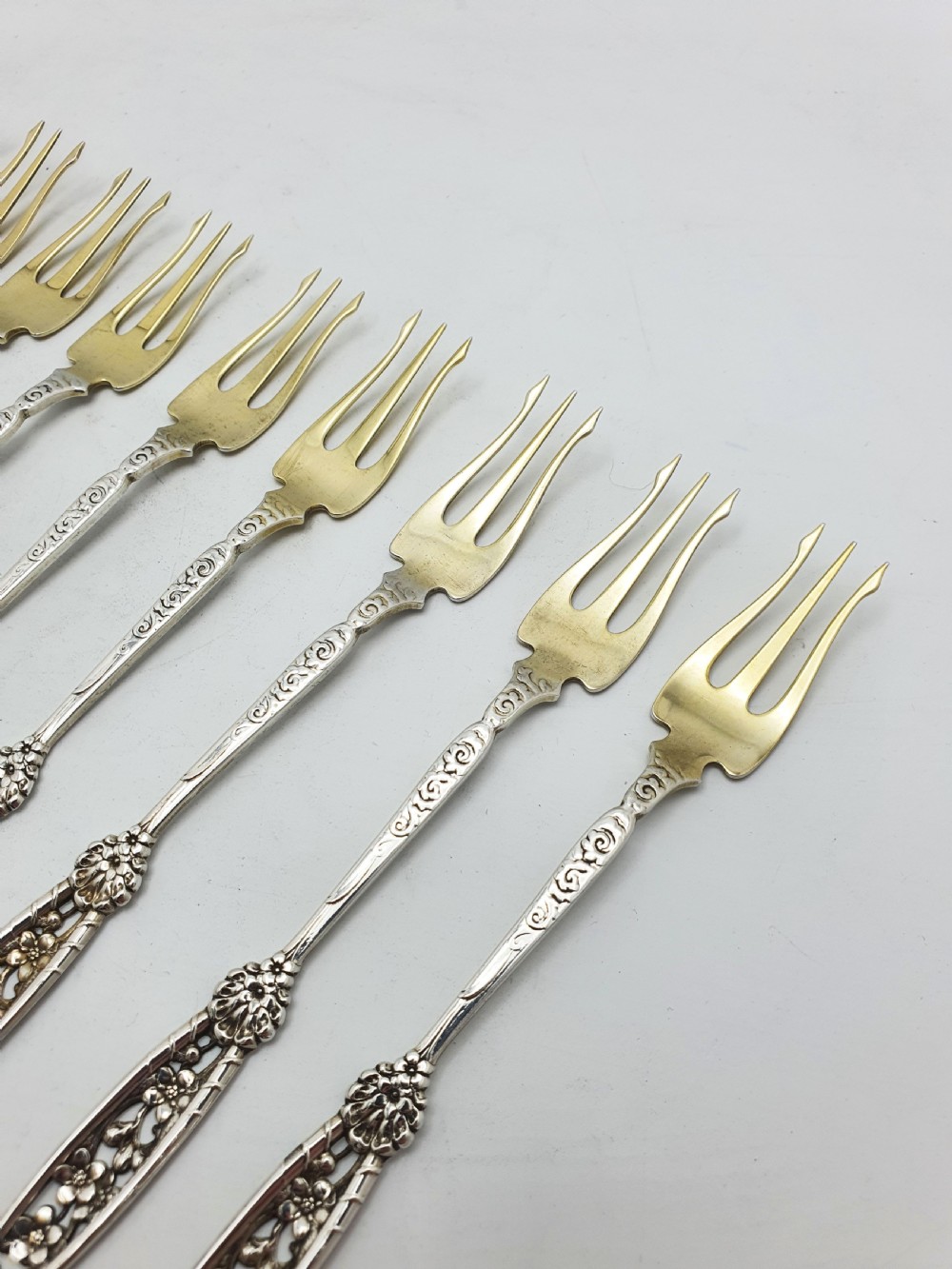 A set of twelve sterling silver seafood forks with ornate floral handles and gold gilt tines arranged in a diagonal line. View V