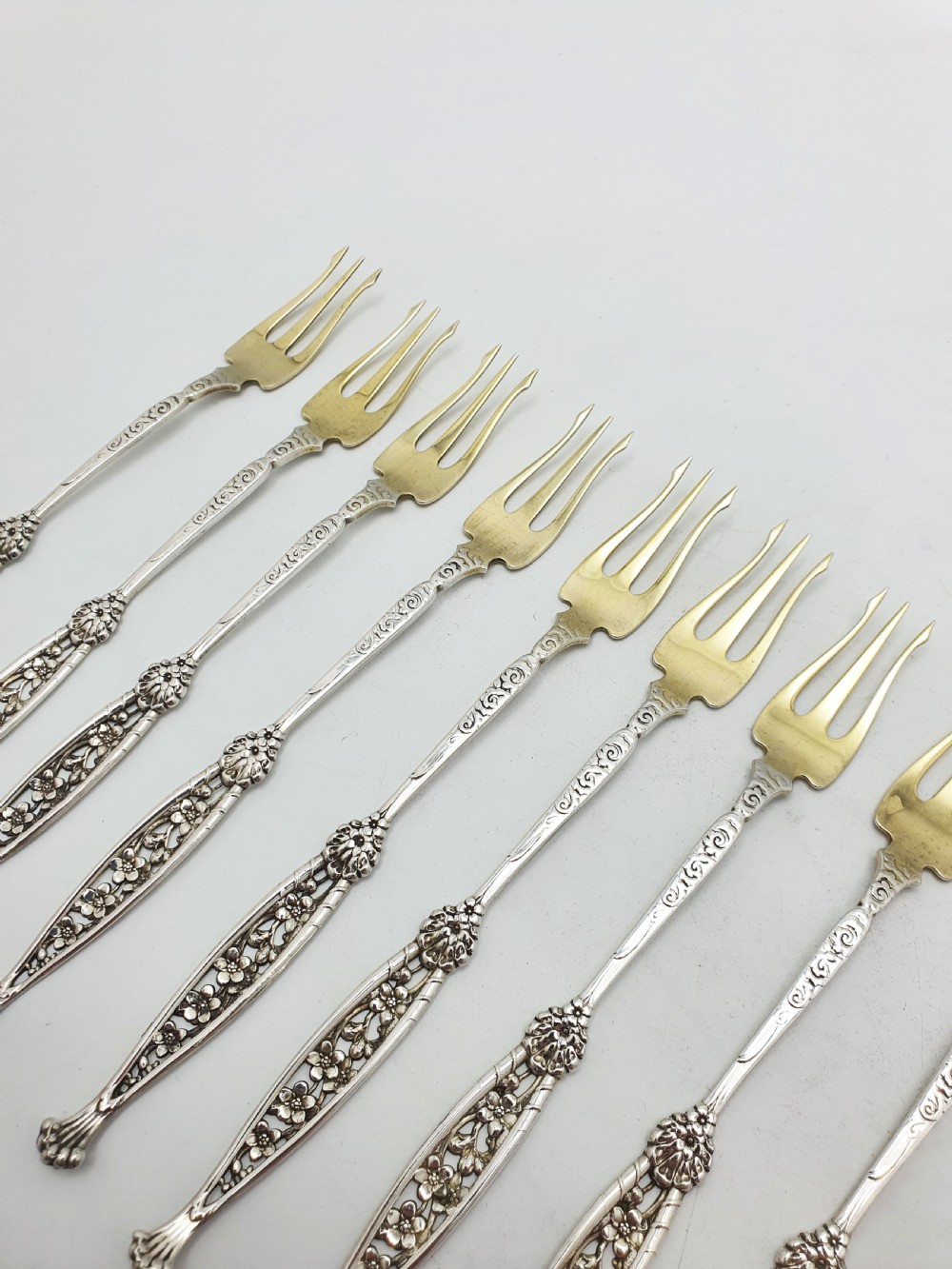 A set of twelve sterling silver seafood forks with ornate floral handles and gold gilt tines arranged in a diagonal line. View VII