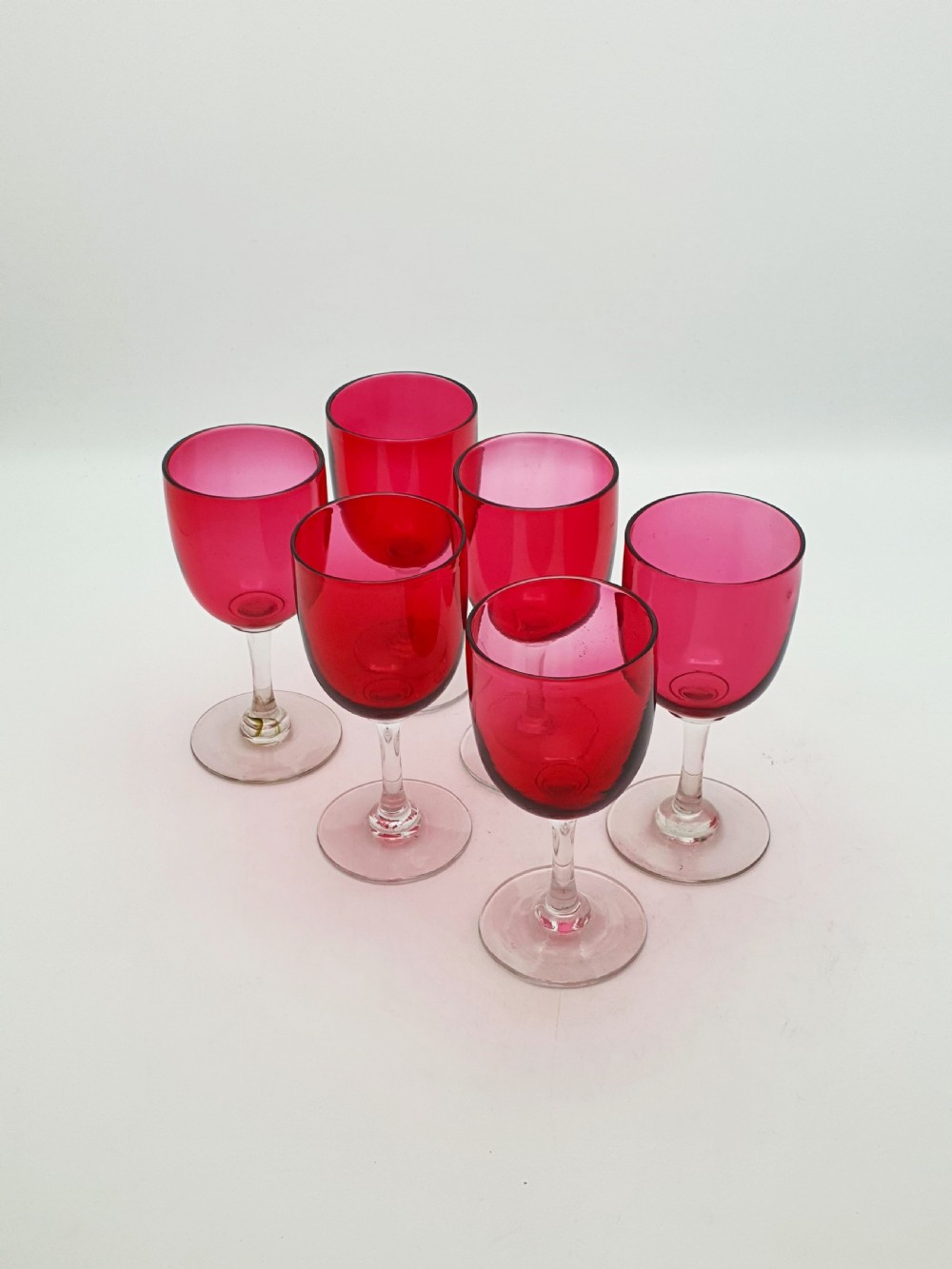 A set of six Victorian hand-blown wine glasses featuring deep cranberry red rounded bowls atop clear glass stems and circular bases.
