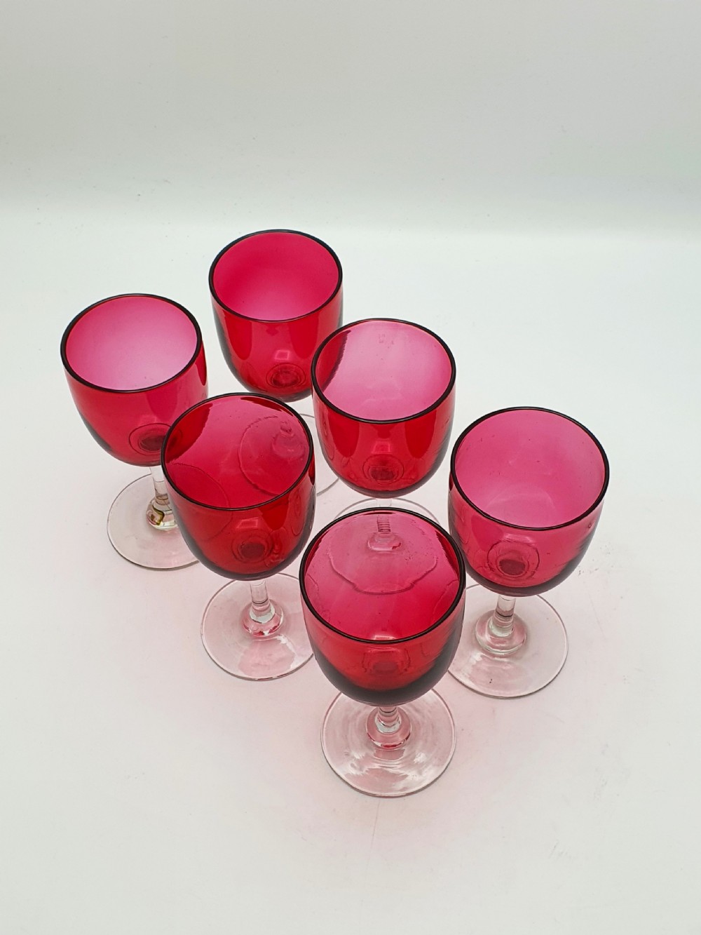 A set of six Victorian hand-blown wine glasses featuring deep cranberry red rounded bowls atop clear glass stems and circular bases. View II