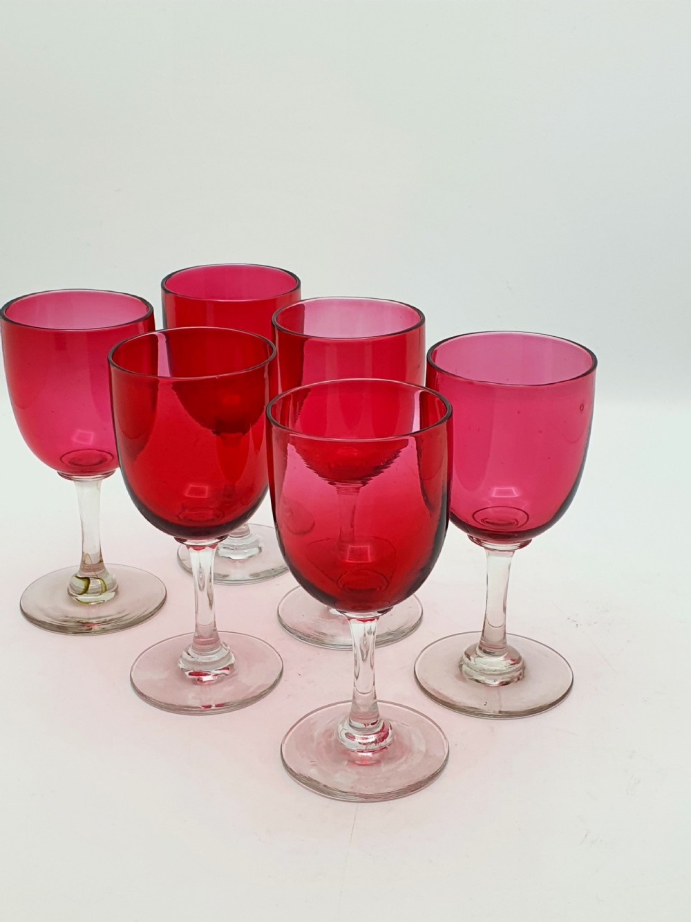 A set of six Victorian hand-blown wine glasses featuring deep cranberry red rounded bowls atop clear glass stems and circular bases. View III