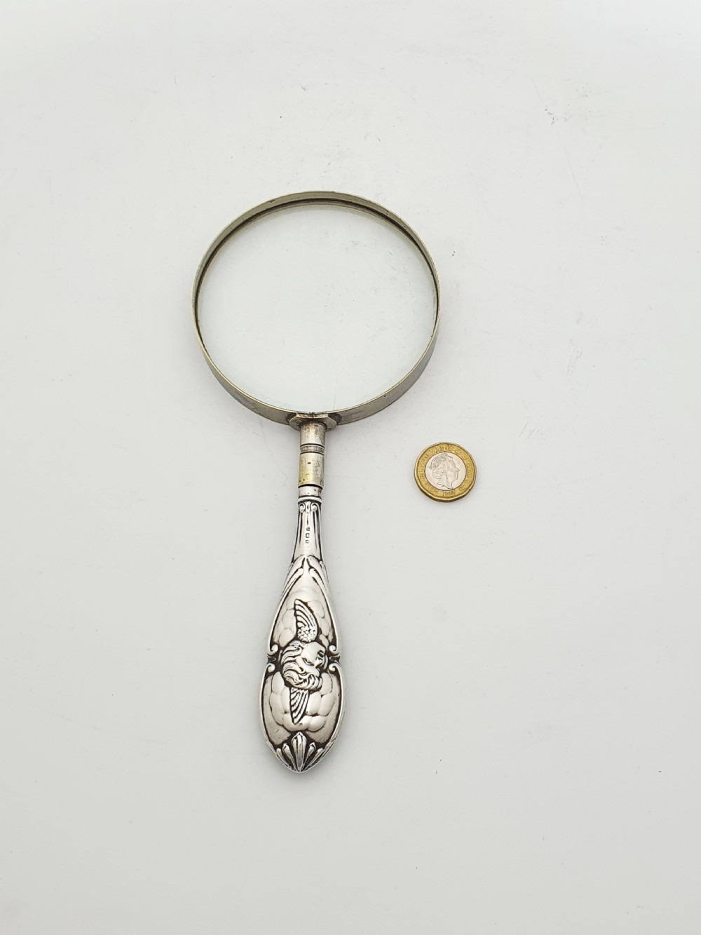An Edwardian magnifying glass featuring a circular glass lens in a metal rim attached to an embossed sterling silver handle. View V