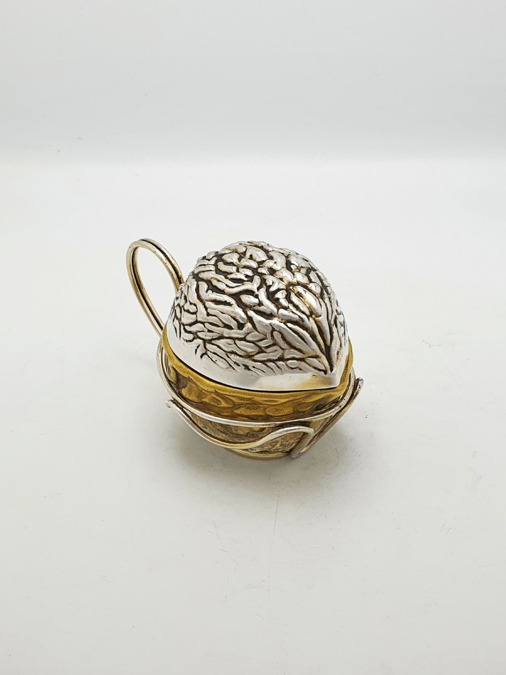 A late Victorian walnut-shaped metal condiment container featuring a textured silver lid and a brass lower shell resting within a wirework stand with a handle.