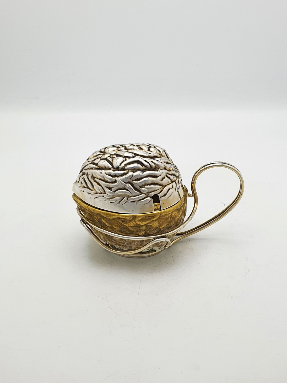 A late Victorian walnut-shaped metal condiment container featuring a textured silver lid and a brass lower shell resting within a wirework stand with a handle. View II