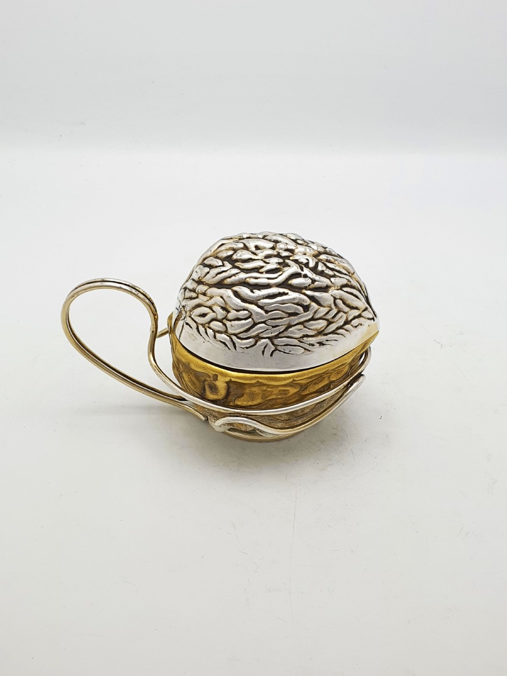 A late Victorian walnut-shaped metal condiment container featuring a textured silver lid and a brass lower shell resting within a wirework stand with a handle. View III