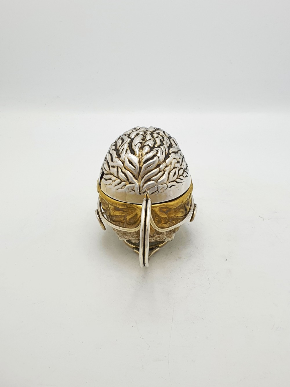 A late Victorian walnut-shaped metal condiment container featuring a textured silver lid and a brass lower shell resting within a wirework stand with a handle. View V