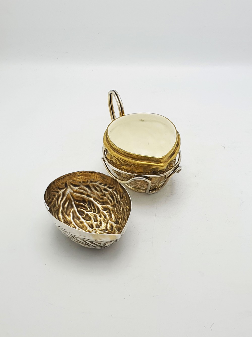 A late Victorian walnut-shaped metal condiment container featuring a textured silver lid and a brass lower shell resting within a wirework stand with a handle. View VI