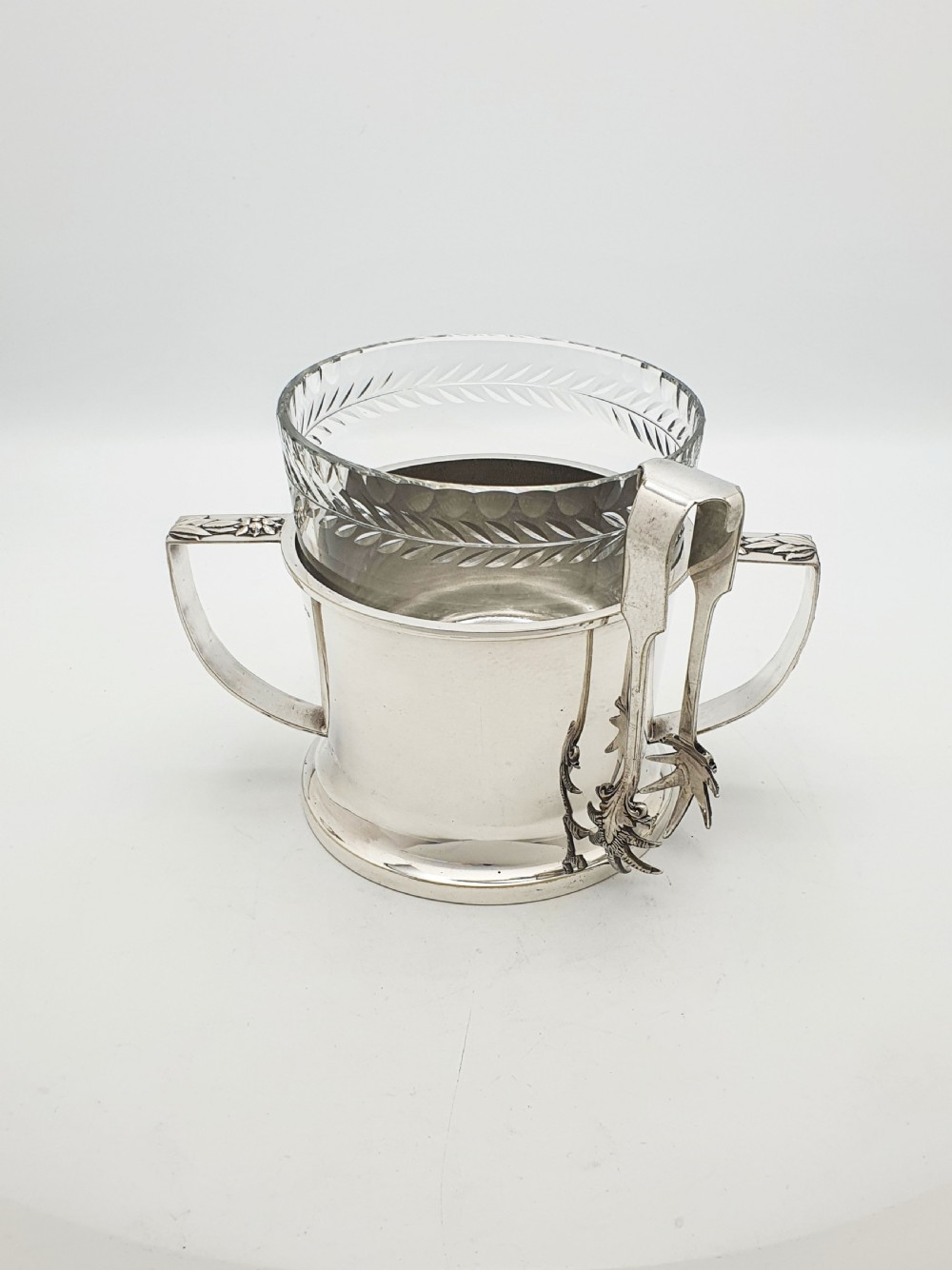 An Art Deco silver-plated ice bucket featuring twin handles, a removable cut-glass liner with a leaf pattern, and matching ice tongs.
