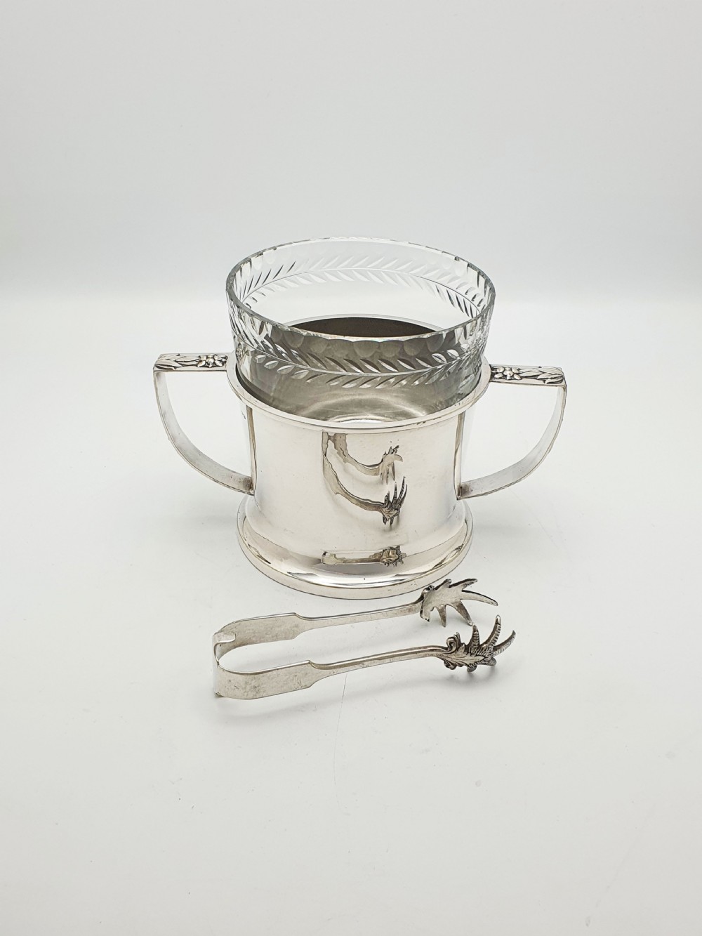 An Art Deco silver-plated ice bucket featuring twin handles, a removable cut-glass liner with a leaf pattern, and matching ice tongs. View II