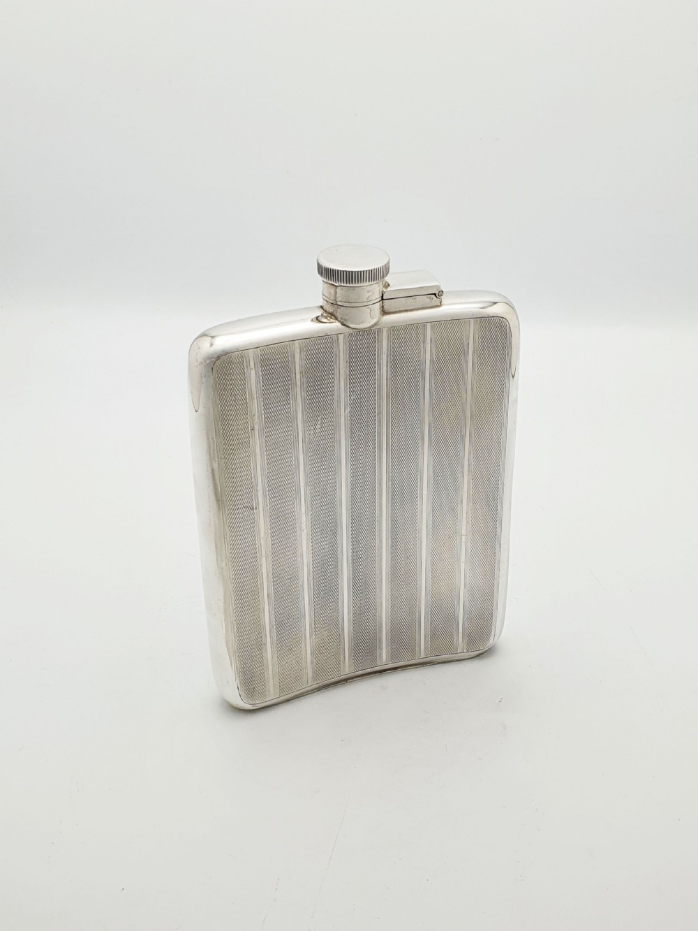 An Art Deco sterling silver hip flask featuring a curved rectangular body decorated with engine-turned linear engraving and a captive bayonet cap.