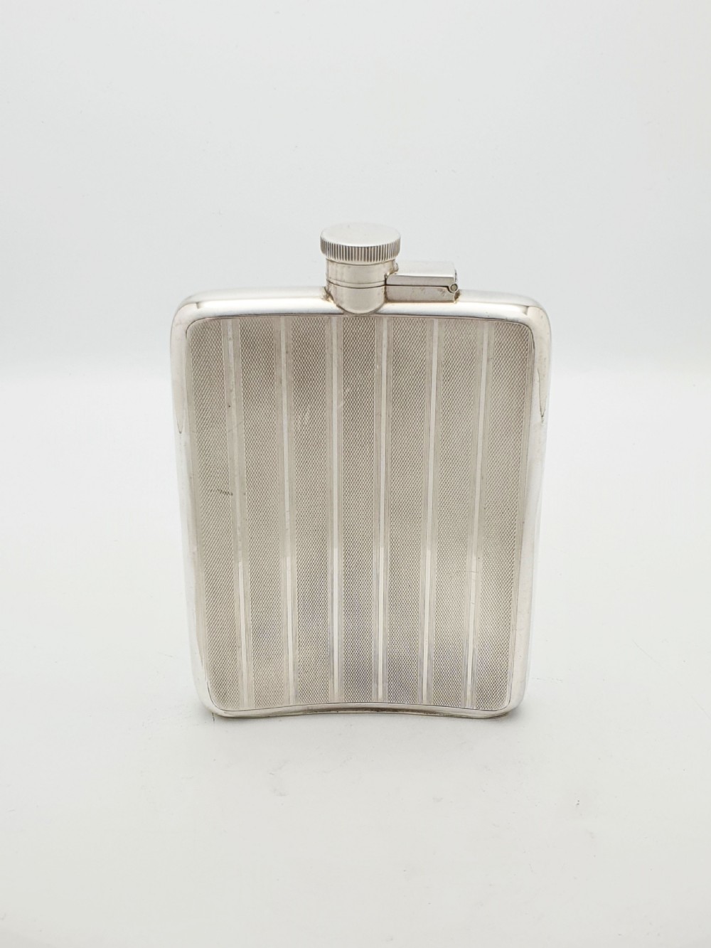 An Art Deco sterling silver hip flask featuring a curved rectangular body decorated with engine-turned linear engraving and a captive bayonet cap. View II