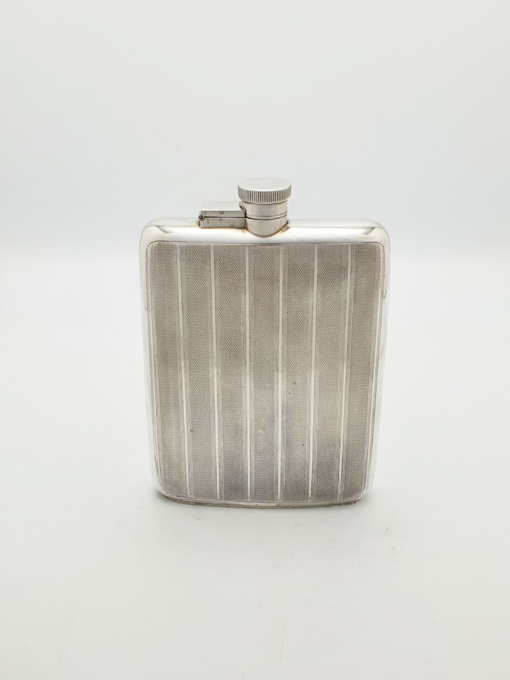 An Art Deco sterling silver hip flask featuring a curved rectangular body decorated with engine-turned linear engraving and a captive bayonet cap. View IV