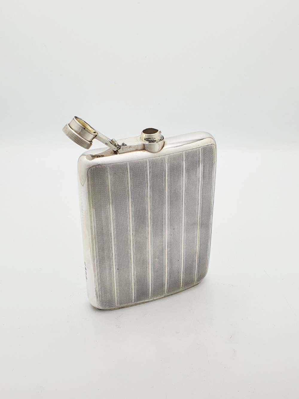 An Art Deco sterling silver hip flask featuring a curved rectangular body decorated with engine-turned linear engraving and a captive bayonet cap. View VI
