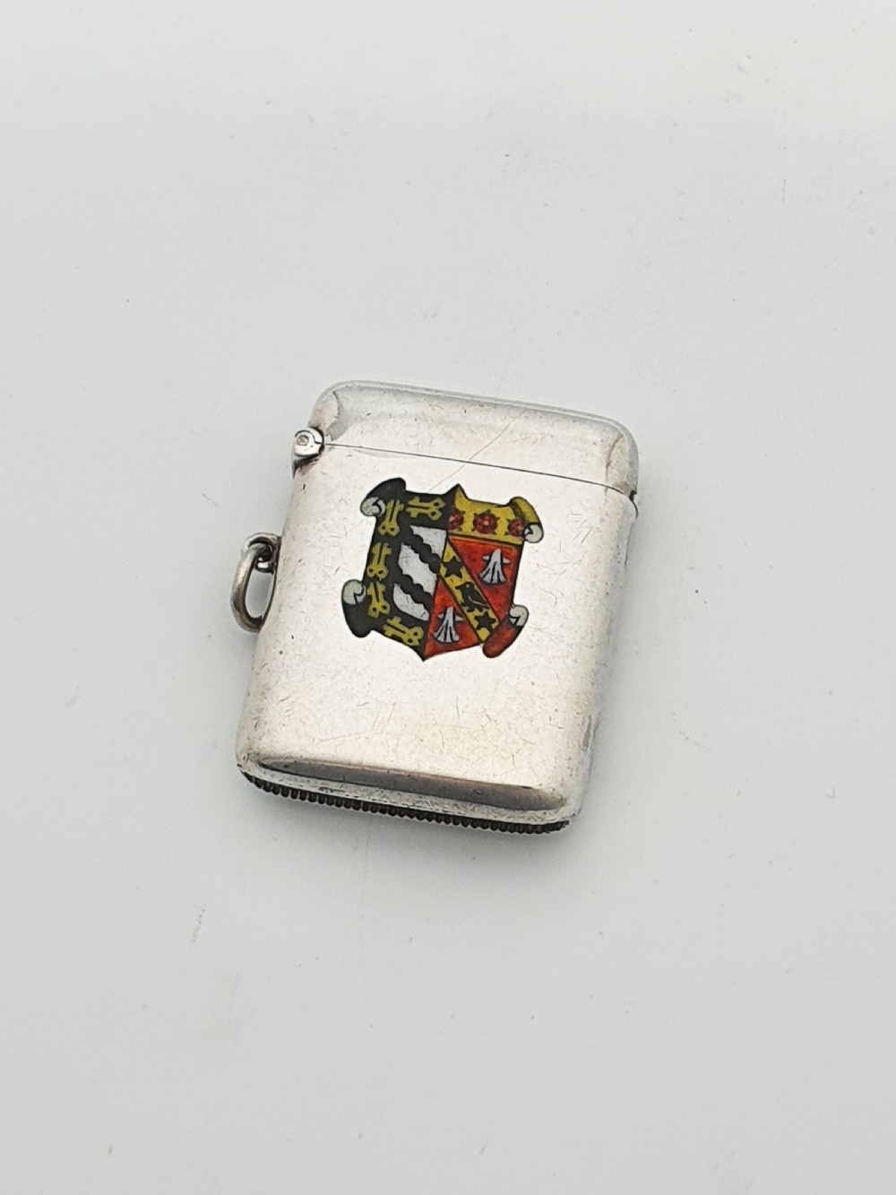 A rectangular sterling silver vesta case featuring a hinged top, side suspension ring, bottom striker, and a colourful heraldic enamel crest on the front.