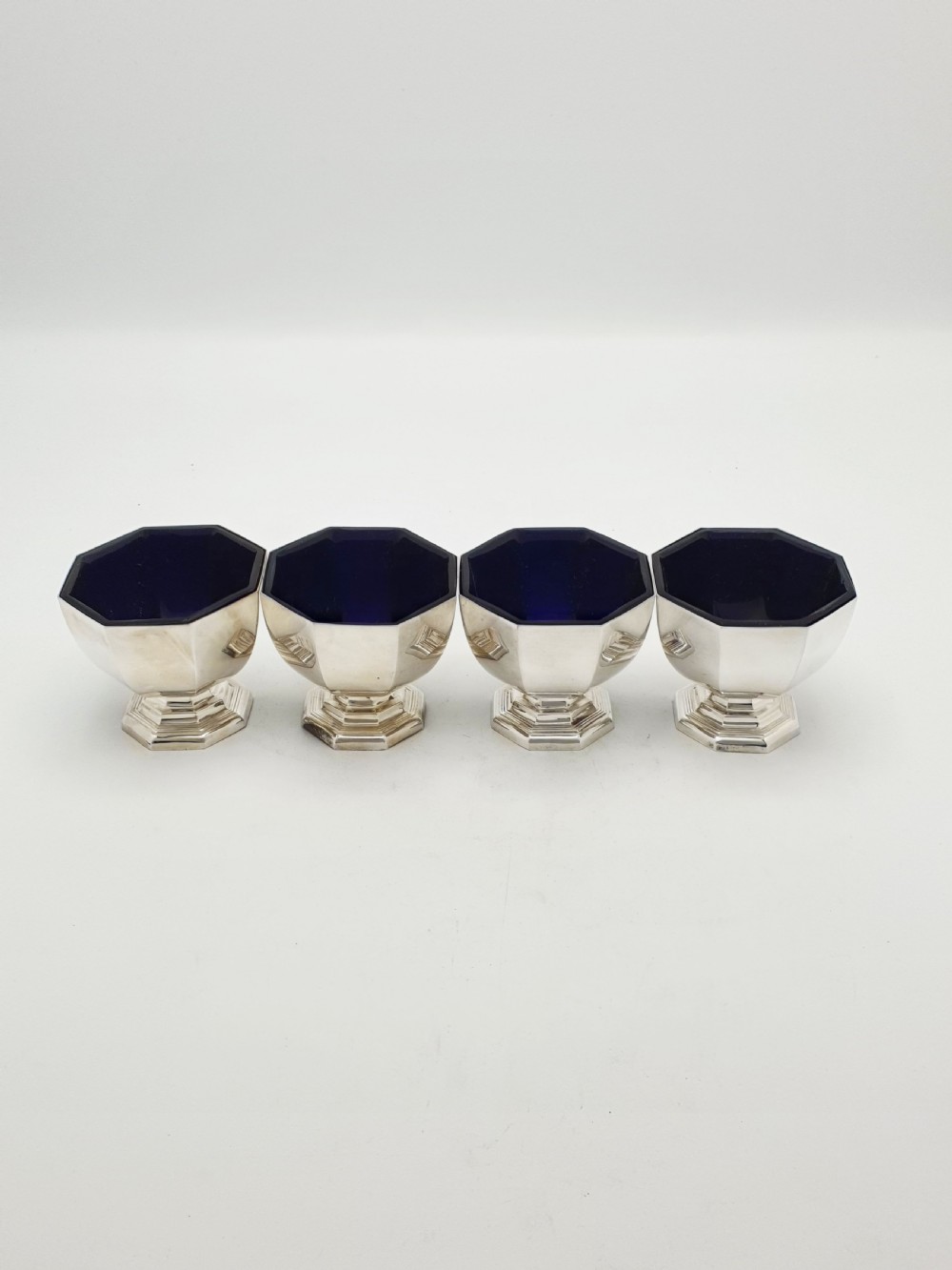 A set of four octagonal silver-plated salt cellars featuring dark cobalt blue glass liners on stepped pedestal bases. View IV
