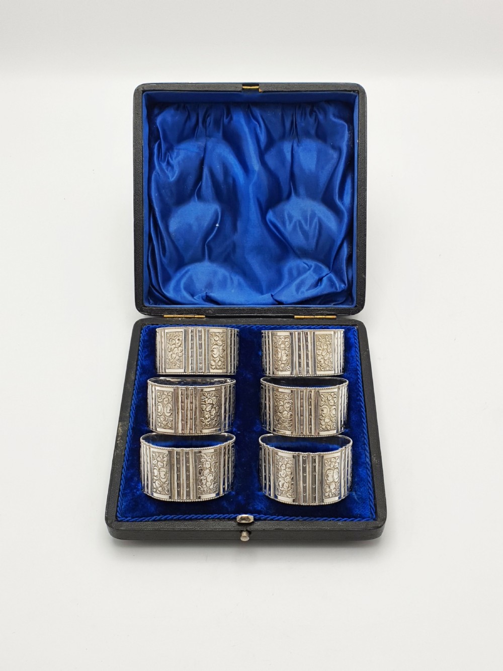 A boxed set of six Edwardian silver-plated napkin rings featuring engraved floral motifs, resting in a blue velvet and silk lined presentation case.