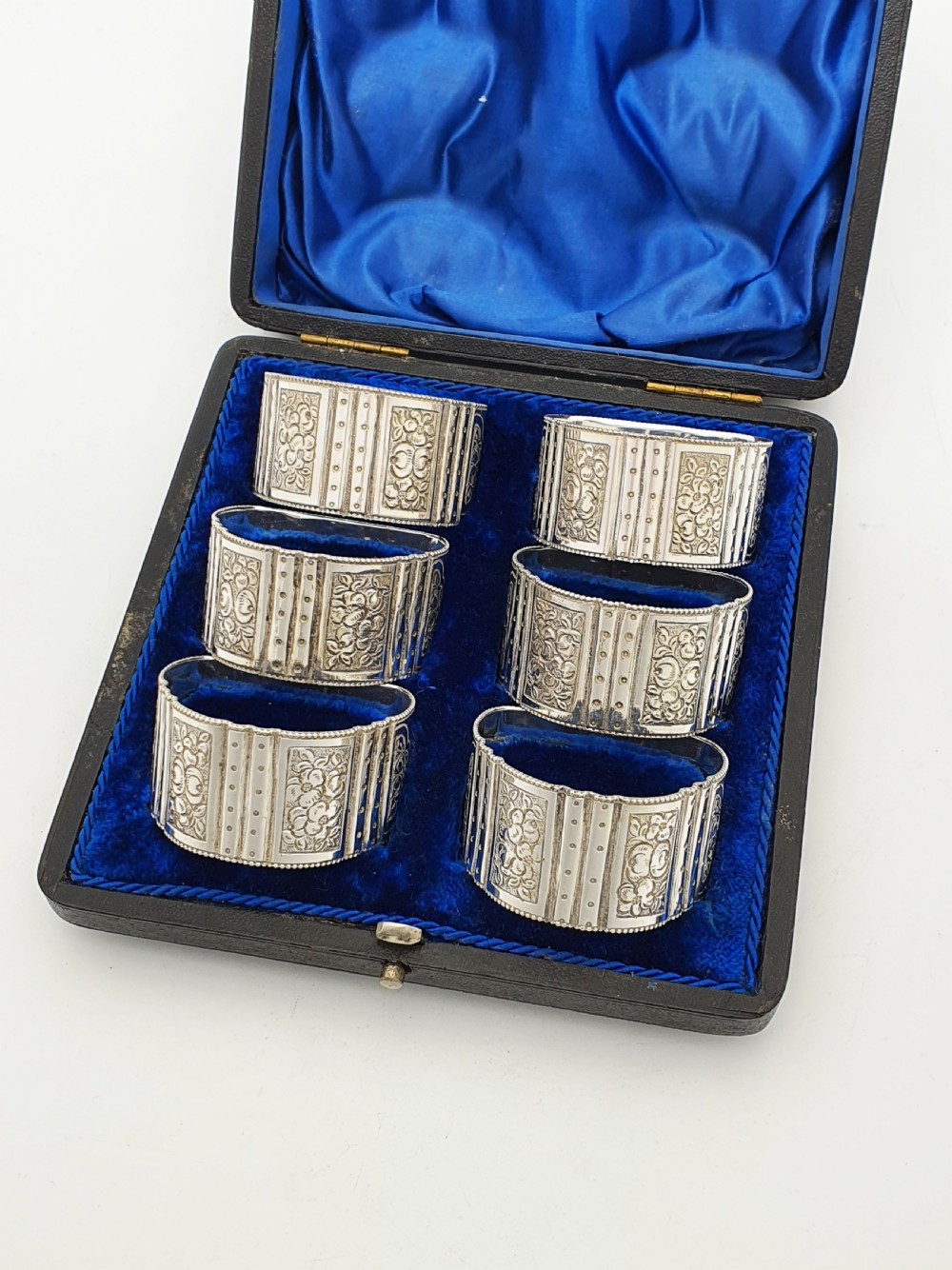 A boxed set of six Edwardian silver-plated napkin rings featuring engraved floral motifs, resting in a blue velvet and silk lined presentation case. View II