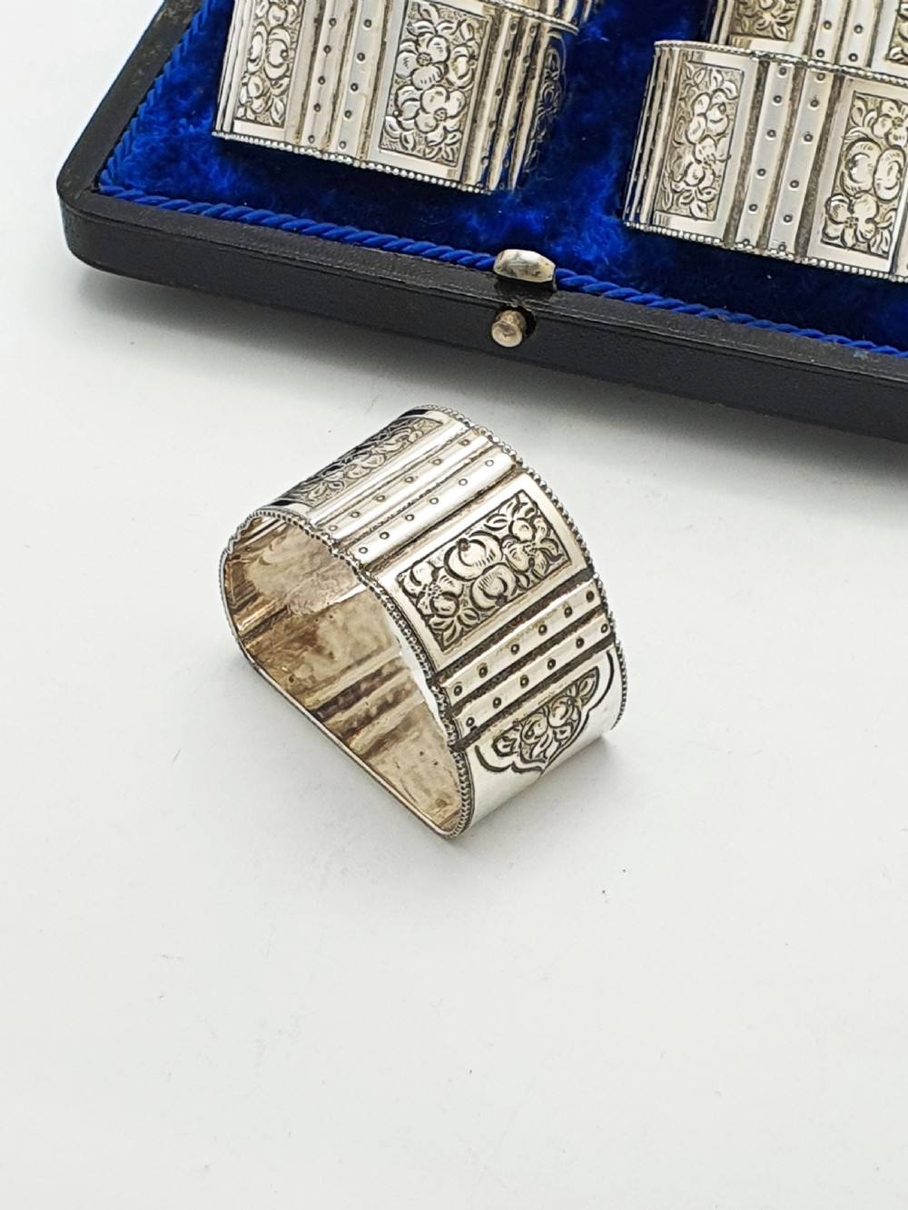 A boxed set of six Edwardian silver-plated napkin rings featuring engraved floral motifs, resting in a blue velvet and silk lined presentation case. View III