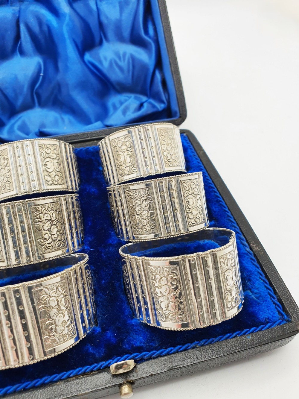 A boxed set of six Edwardian silver-plated napkin rings featuring engraved floral motifs, resting in a blue velvet and silk lined presentation case. View IV