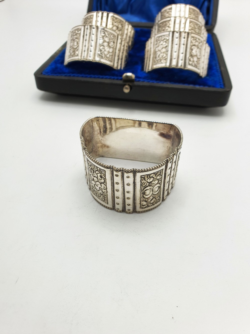 A boxed set of six Edwardian silver-plated napkin rings featuring engraved floral motifs, resting in a blue velvet and silk lined presentation case. View V