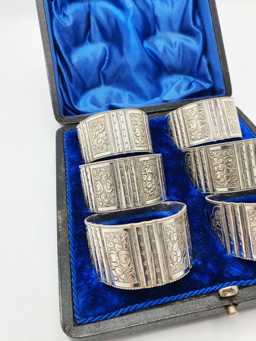 A boxed set of six Edwardian silver-plated napkin rings featuring engraved floral motifs, resting in a blue velvet and silk lined presentation case. View VI