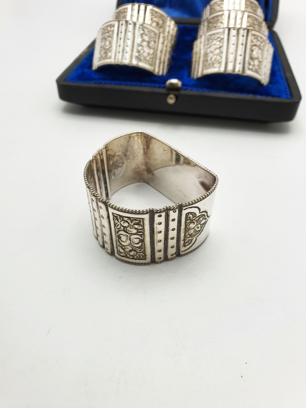 A boxed set of six Edwardian silver-plated napkin rings featuring engraved floral motifs, resting in a blue velvet and silk lined presentation case. View VII