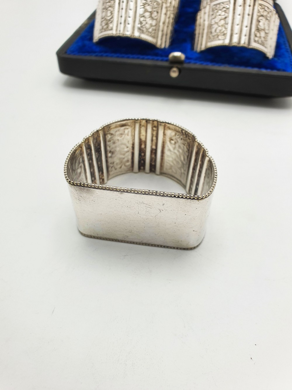 A boxed set of six Edwardian silver-plated napkin rings featuring engraved floral motifs, resting in a blue velvet and silk lined presentation case. View VIII