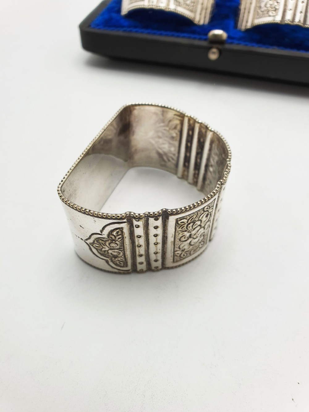 A boxed set of six Edwardian silver-plated napkin rings featuring engraved floral motifs, resting in a blue velvet and silk lined presentation case. View IX