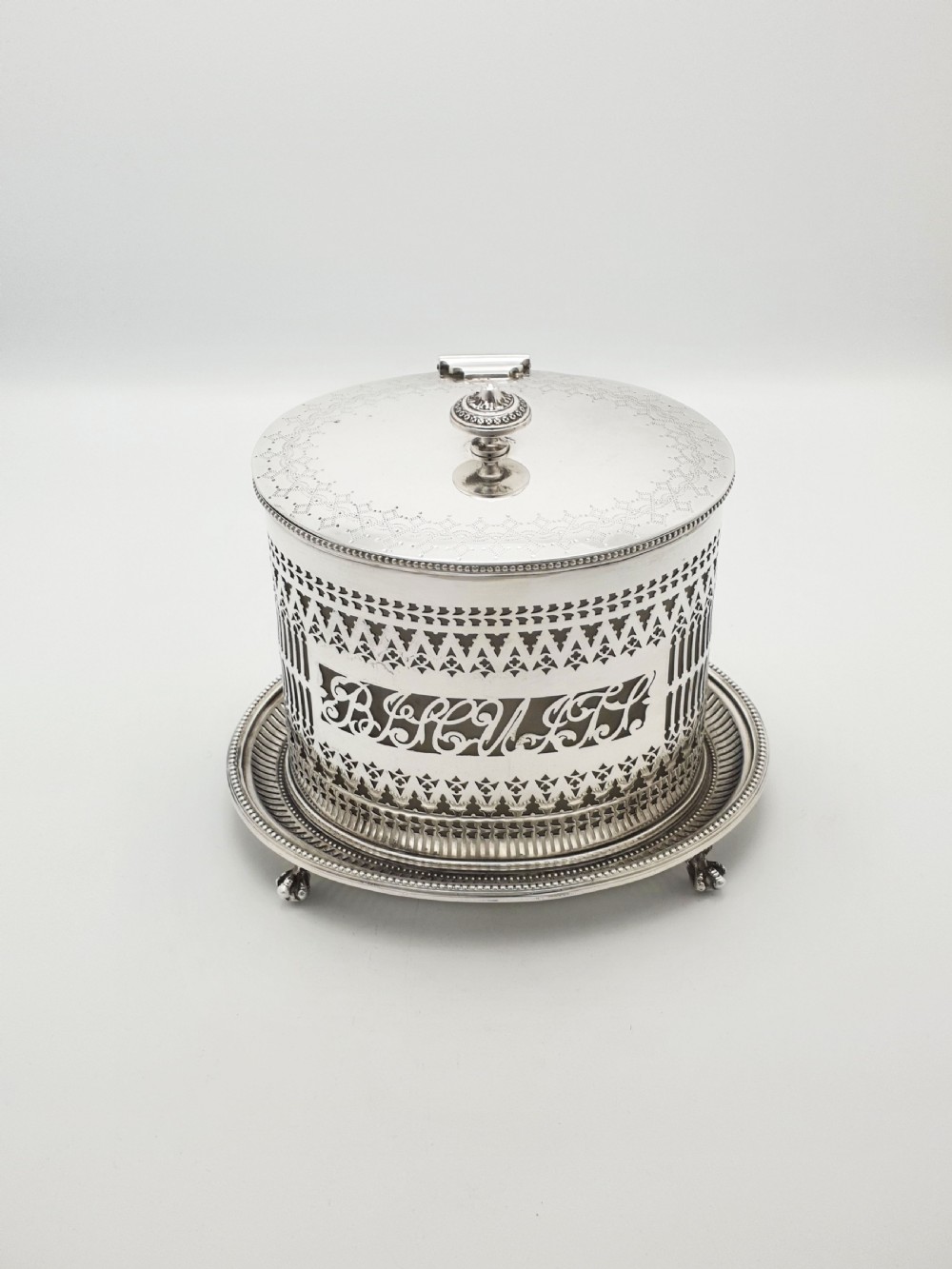 An Edwardian silver plated biscuit barrel featuring pierced scrollwork spelling out the word biscuits, a matching tray base, and a hinged lid with a decorative finial.