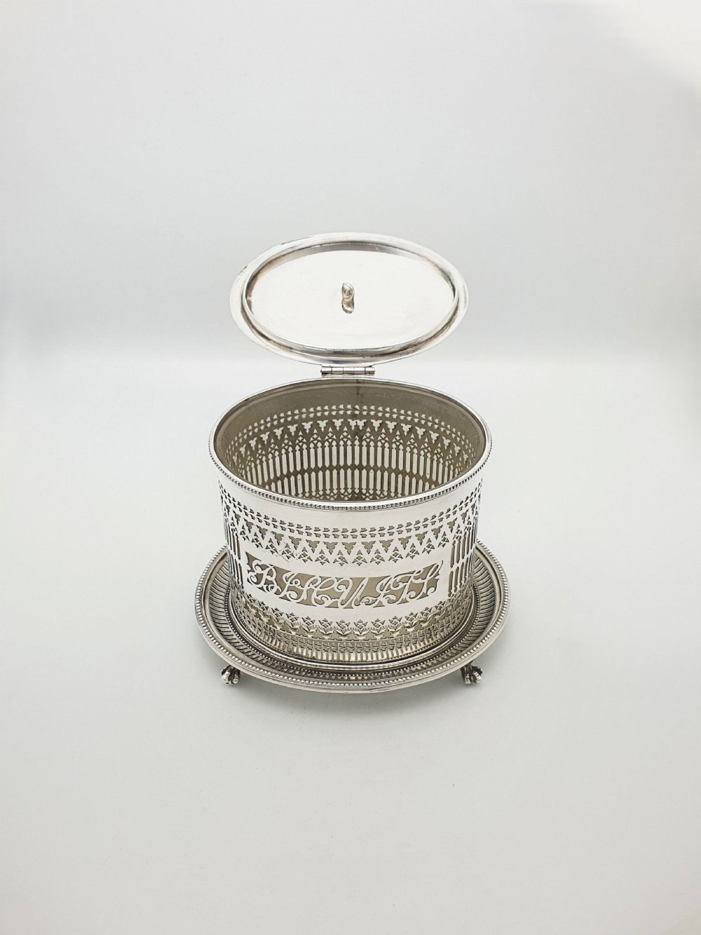 An Edwardian silver plated biscuit barrel featuring pierced scrollwork spelling out the word biscuits, a matching tray base, and a hinged lid with a decorative finial. View II