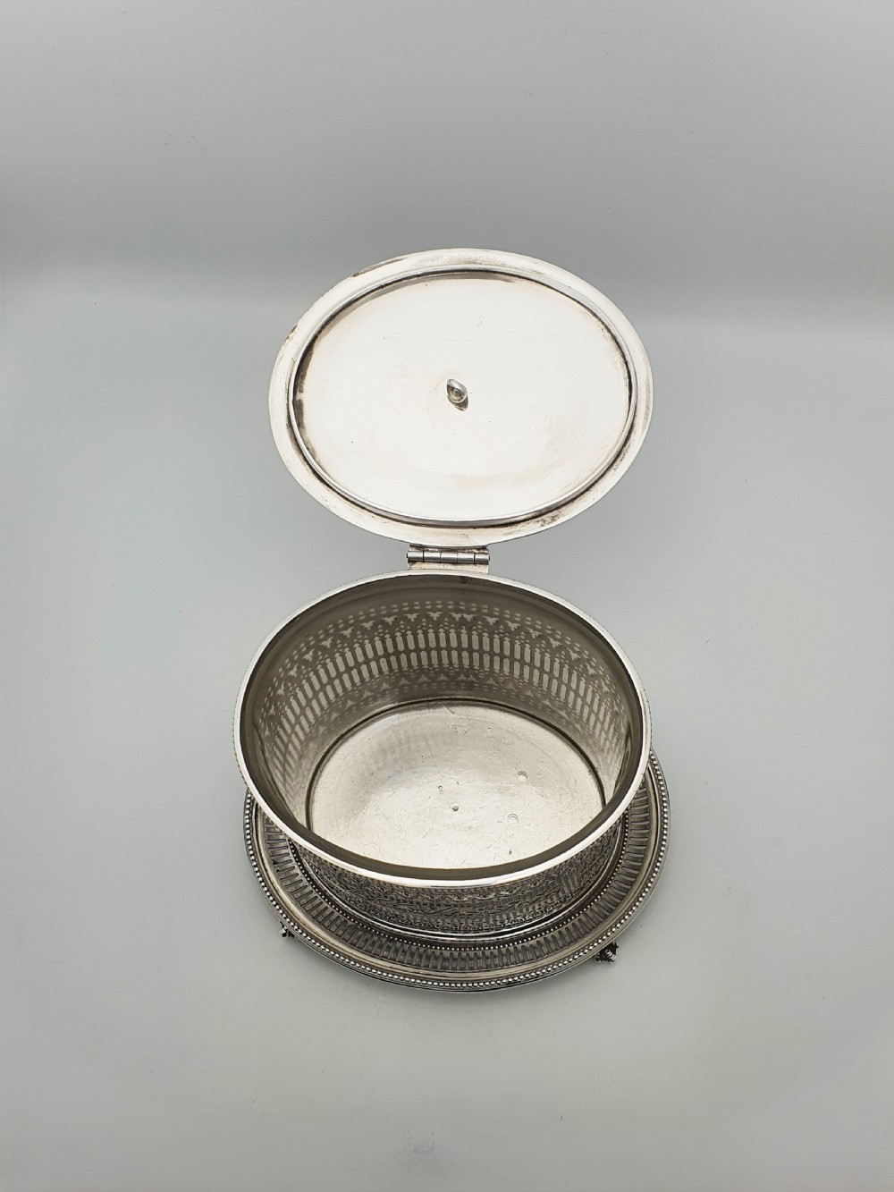 An Edwardian silver plated biscuit barrel featuring pierced scrollwork spelling out the word biscuits, a matching tray base, and a hinged lid with a decorative finial. View III